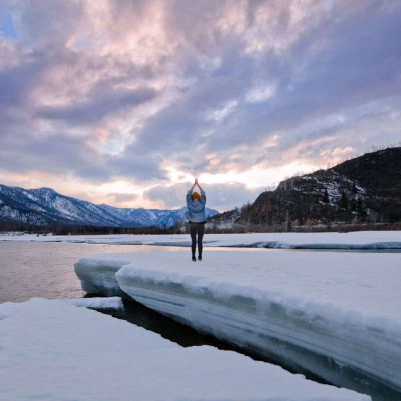 A woman with raised arms stands on an ice floe by a lake amidst snowy hills at dusk.