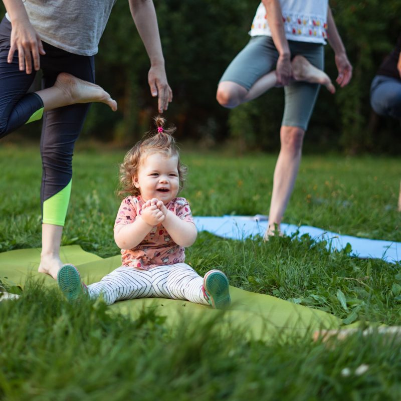 A cheerful toddler sits on a yoga mat surrounded by adults practicing yoga in a sunny park.