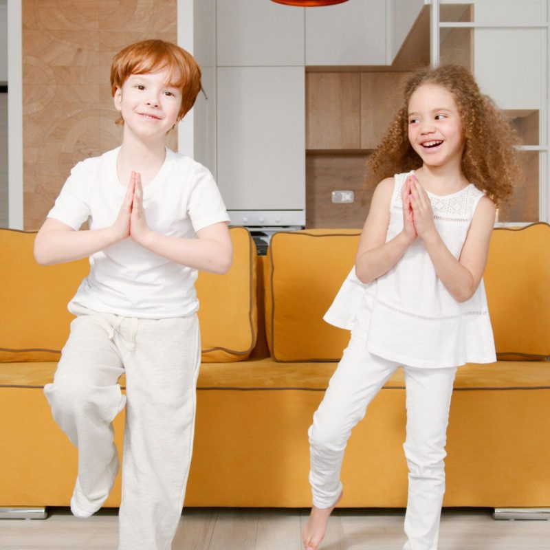 Two kids practicing yoga indoors, standing in tree pose with smiles, showcasing a sense of balance and enjoyment.
